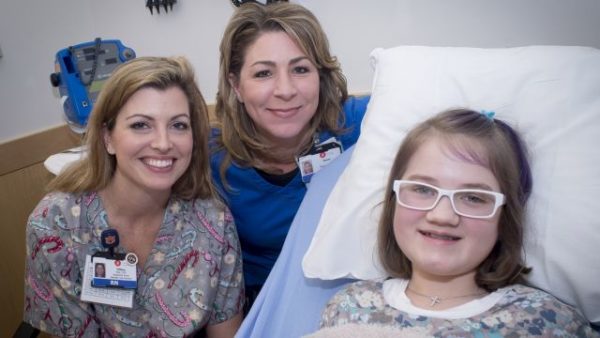 Two nurses are sitting beside Mandalynn Smith (Pediatric Patient) who is laying in a hospital bed at Children's Hospital of Alabama before an infusion of pamidronate, a drug in the bisphosphonate family, used to treat bone diseases, as part of UAB's Undiagnosed Diseases Program, 2015.